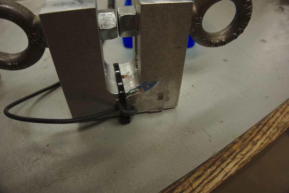 A terribly framed picture of the final hardware. You pull on the two eyebolts at the top of the photo, and the inside elbow of the U stretches. The two little brown tabs are the strain gauges. The one in the elbow actually measures the stretch, and the other on the side experiences no stretch and thus acts as a baseline for correcting temperature change. The cable attaching the gauges to the circuitry is held in place with a zip-tie to prevent the strain gauges from getting ripped off accidentally.